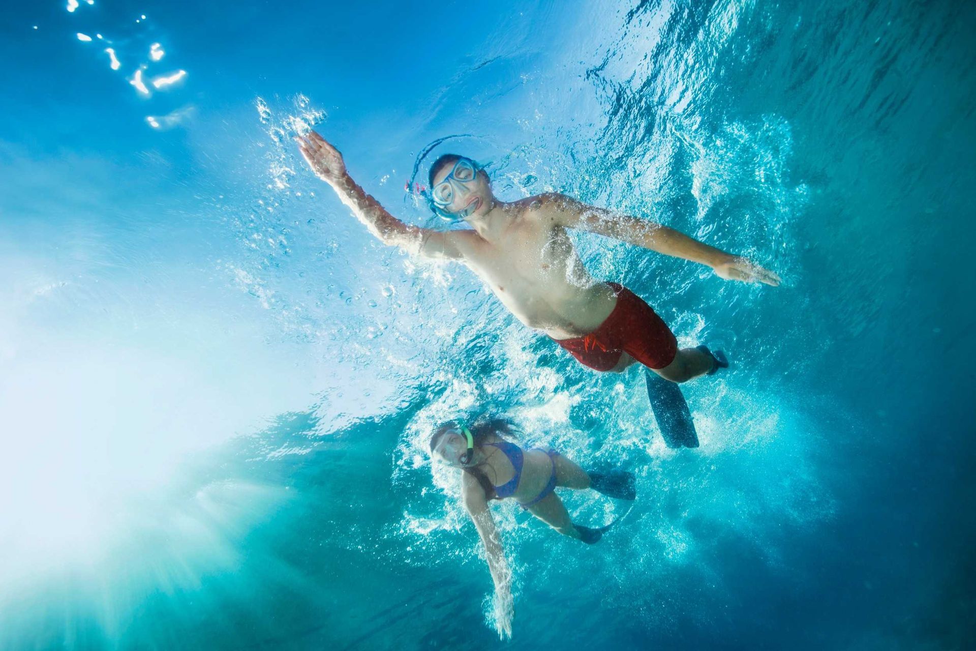 Two swimmers underwater in a bright blue ocean, reaching through a wave