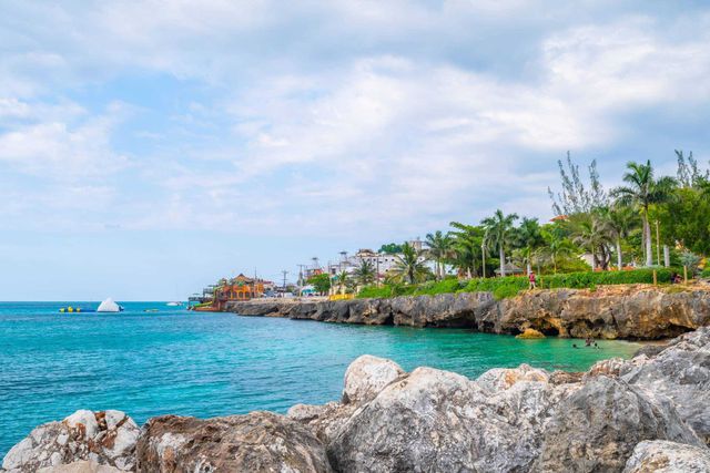 Rocky tropical shoreline with turquoise water, palm trees, and a cloudy blue sky