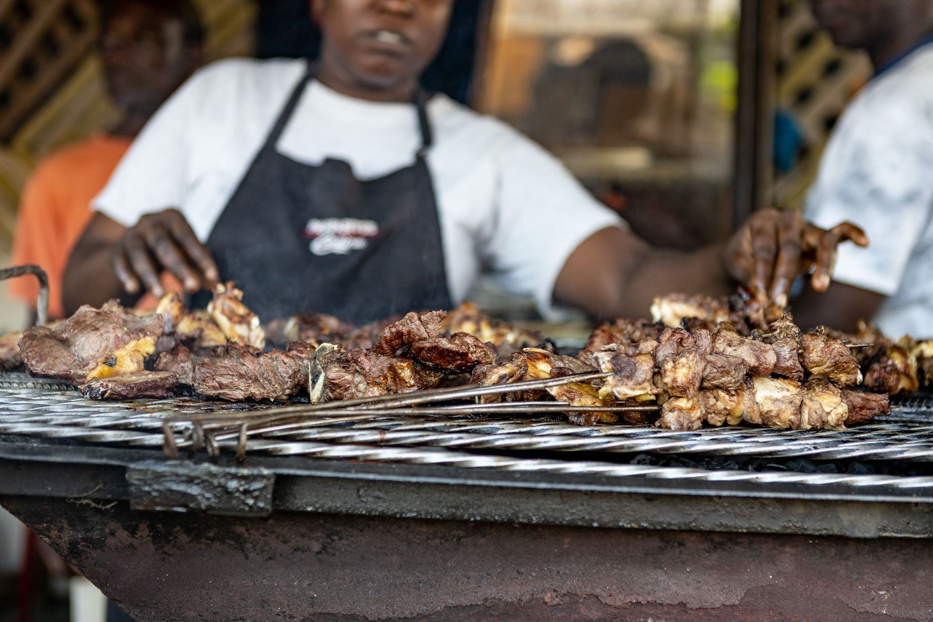 Grilled meat sizzling on a barbecue grill, with a cook in apron tending the food