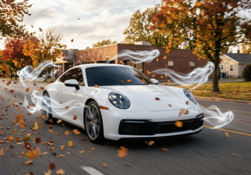 White Porsche 911 driving along a tree-lined road surrounded by falling autumn leaves and ghosts