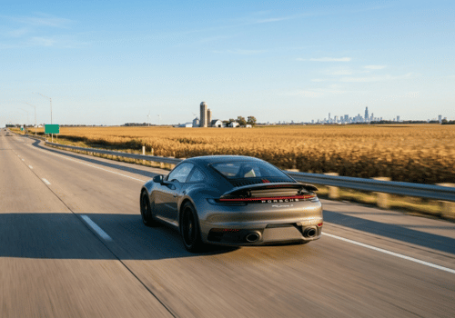 Porsche sports car driving on a highway through open farmland with a distant city skyline on the horizon.