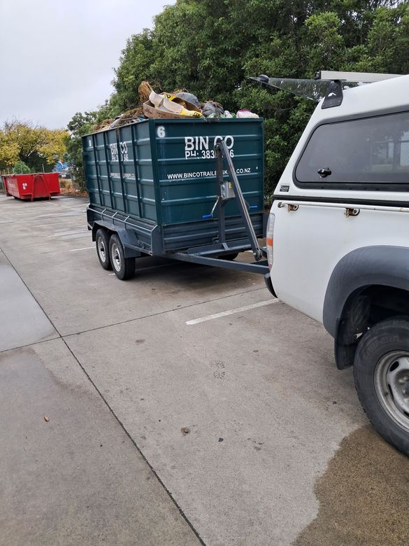 A white pickup truck is parked in a lot, towing a dark green waste skip bin filled with junk.