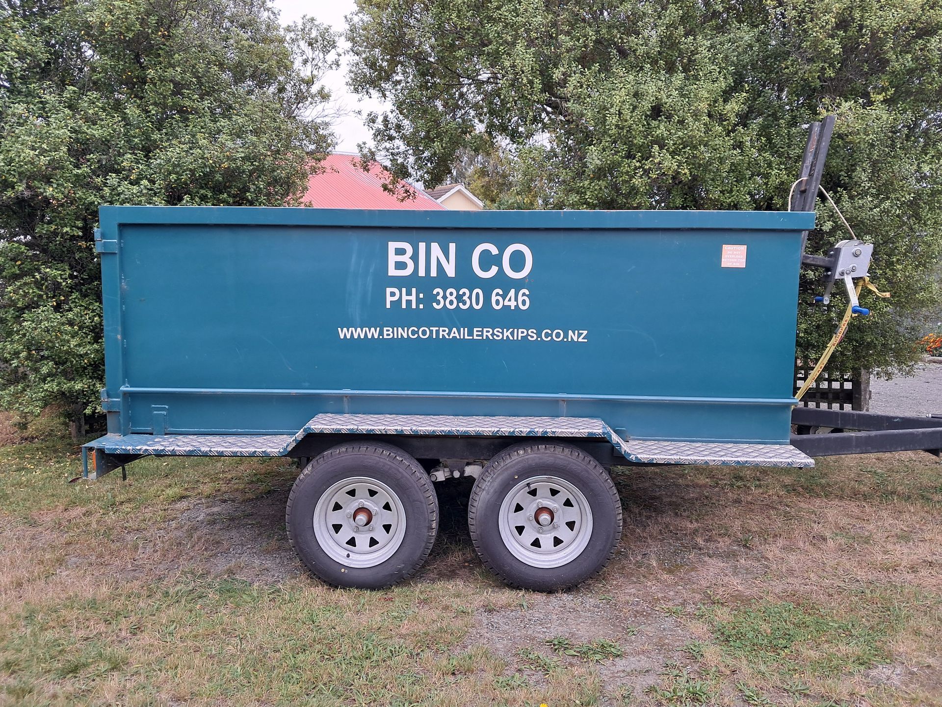 A teal trailer with the text BIN CO, a phone number, and a website, parked on grass in front of trees.