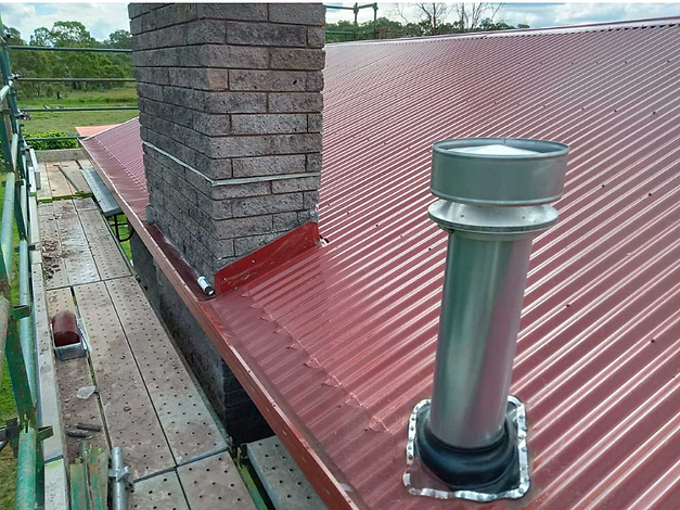 Red corrugated metal roof with a brick chimney and silver vent pipe beside a scaffolded edge