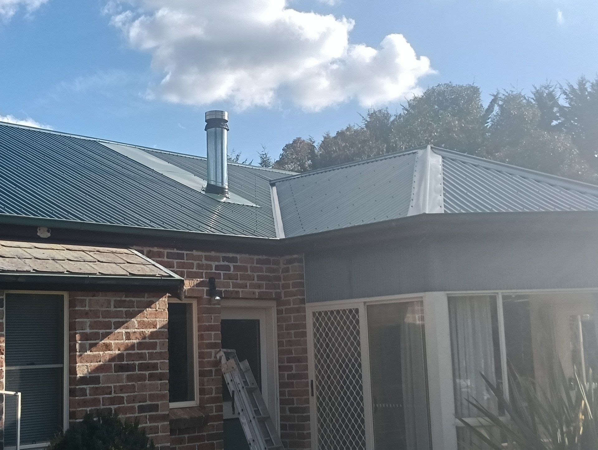 Red corrugated metal roof with a brick chimney and silver vent pipe beside a scaffolded edge