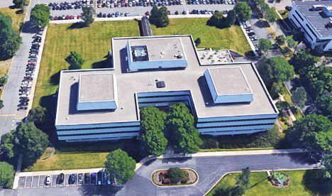 Aerial view of a multi-story office building surrounded by green lawn, trees, and parking lots.