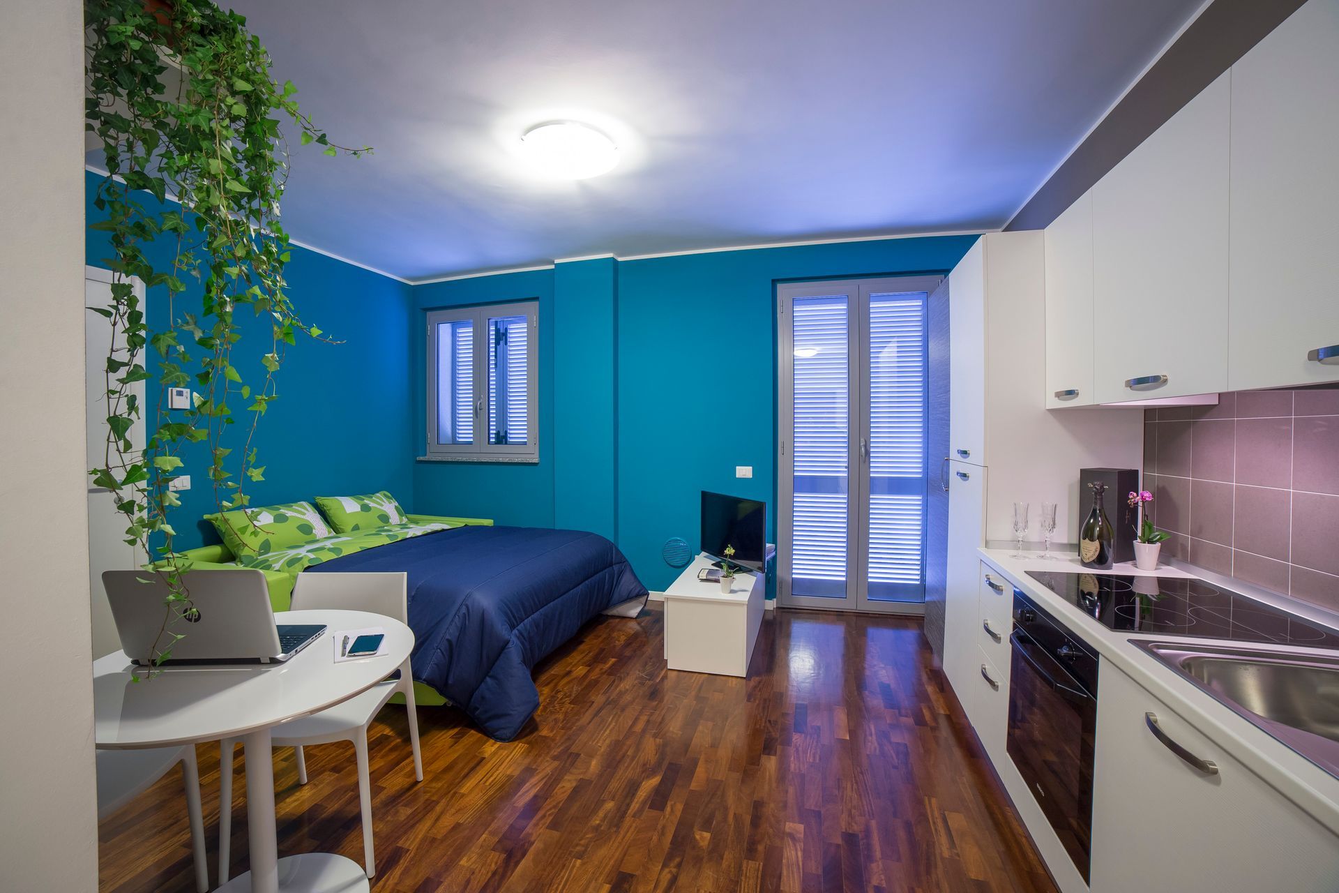 Piccolo monolocale con pareti turchesi, cucina bianca, letto e tavolo rotondo.