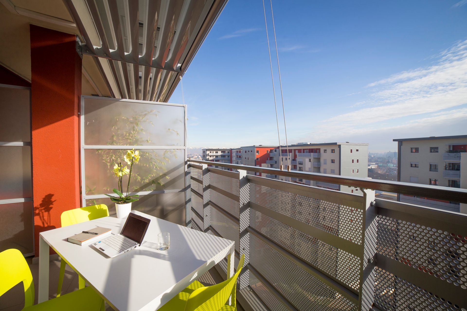Balcone con tavolo bianco, computer portatile e sedie gialle, con vista sugli edifici della città in una giornata di sole.