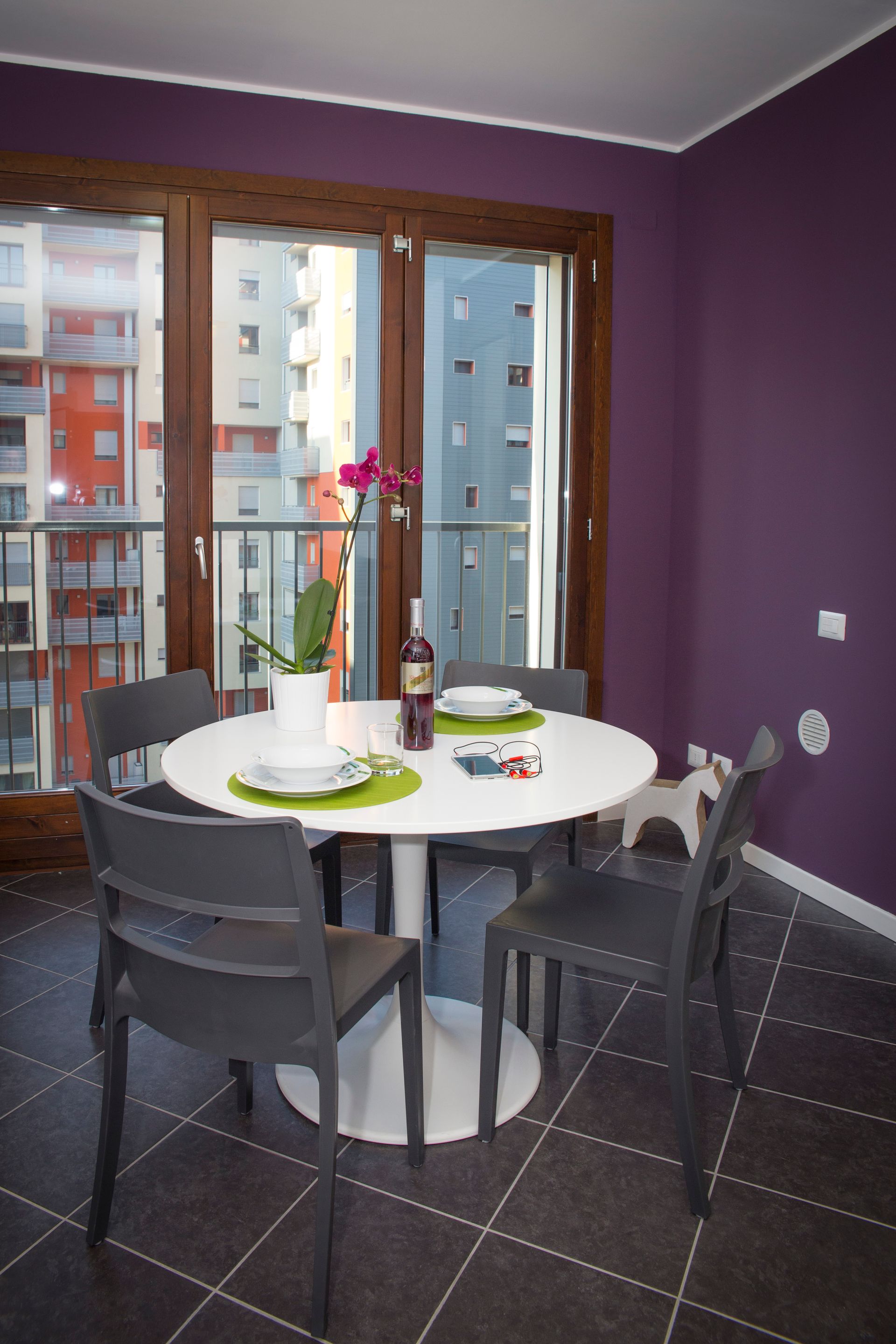 Sala da pranzo con tavolo rotondo bianco, quattro sedie grigie, pareti viola e vista sulla città.