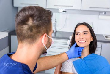 A dentist in blue scrubs and a mask touches the chin of a smiling patient in a clinical dental office.
