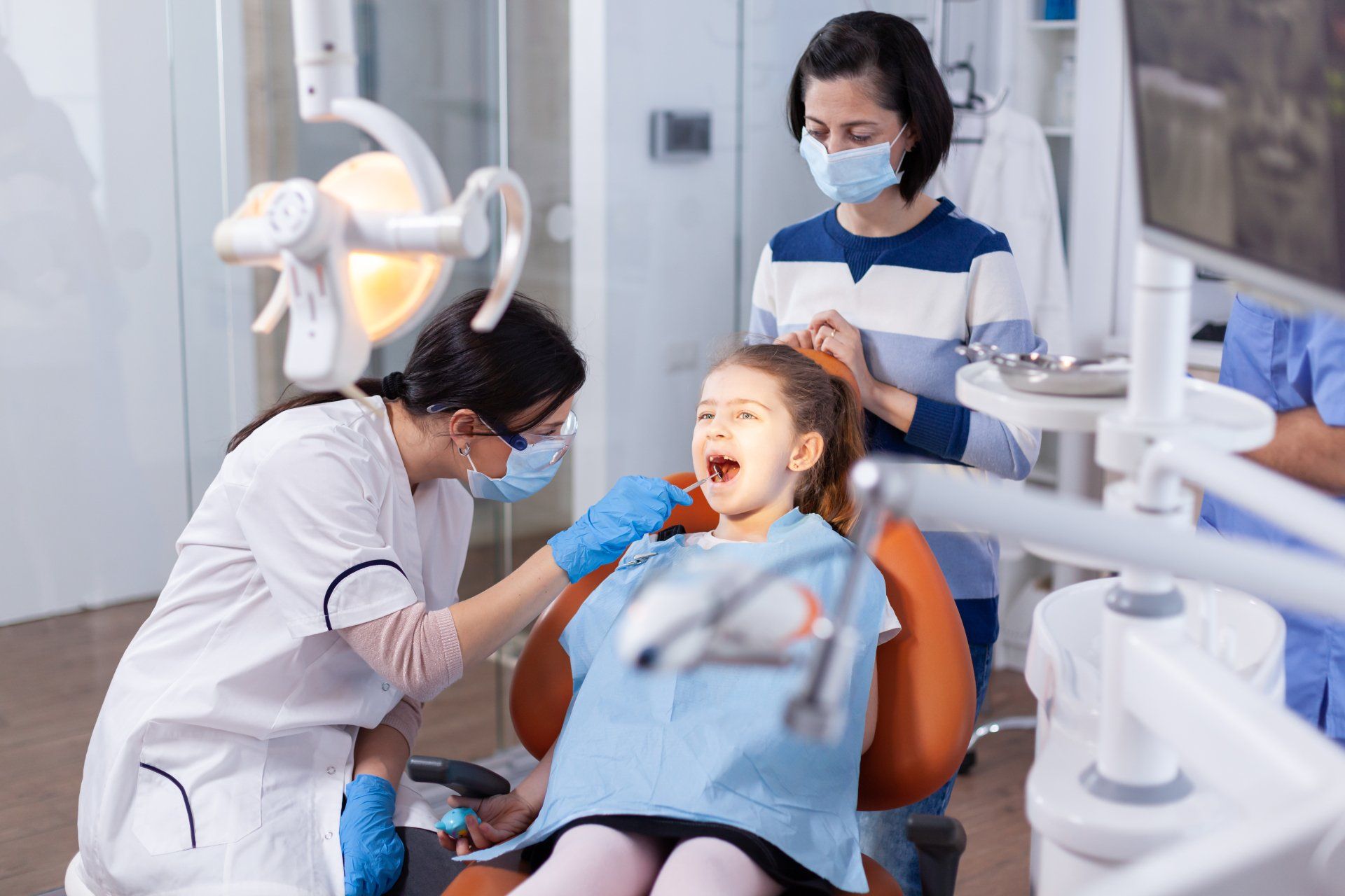 A dentist performs an examination on a child in a dental office while an adult watches nearby.