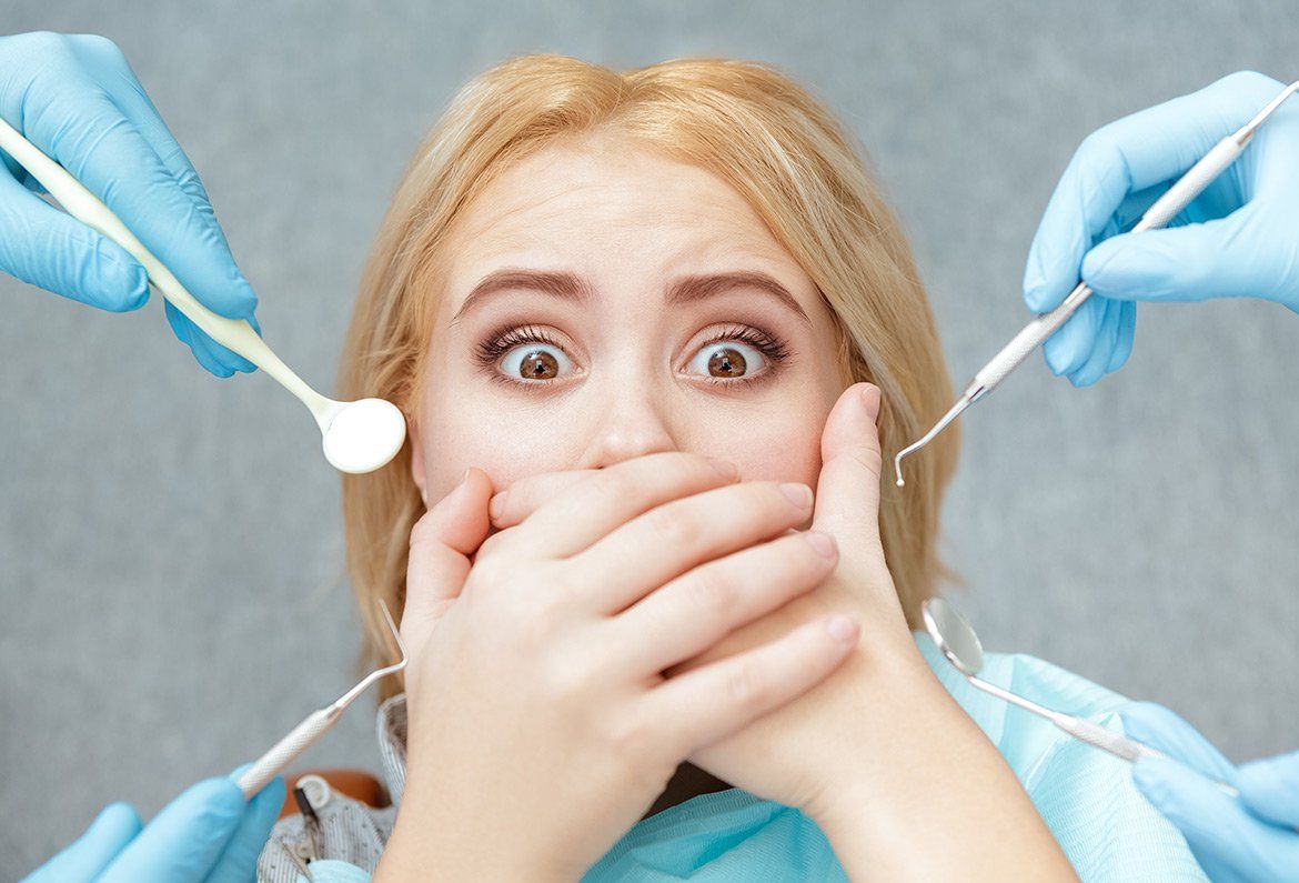 A person with a startled expression covers their mouth with their hands, surrounded by hands holding dental instruments.