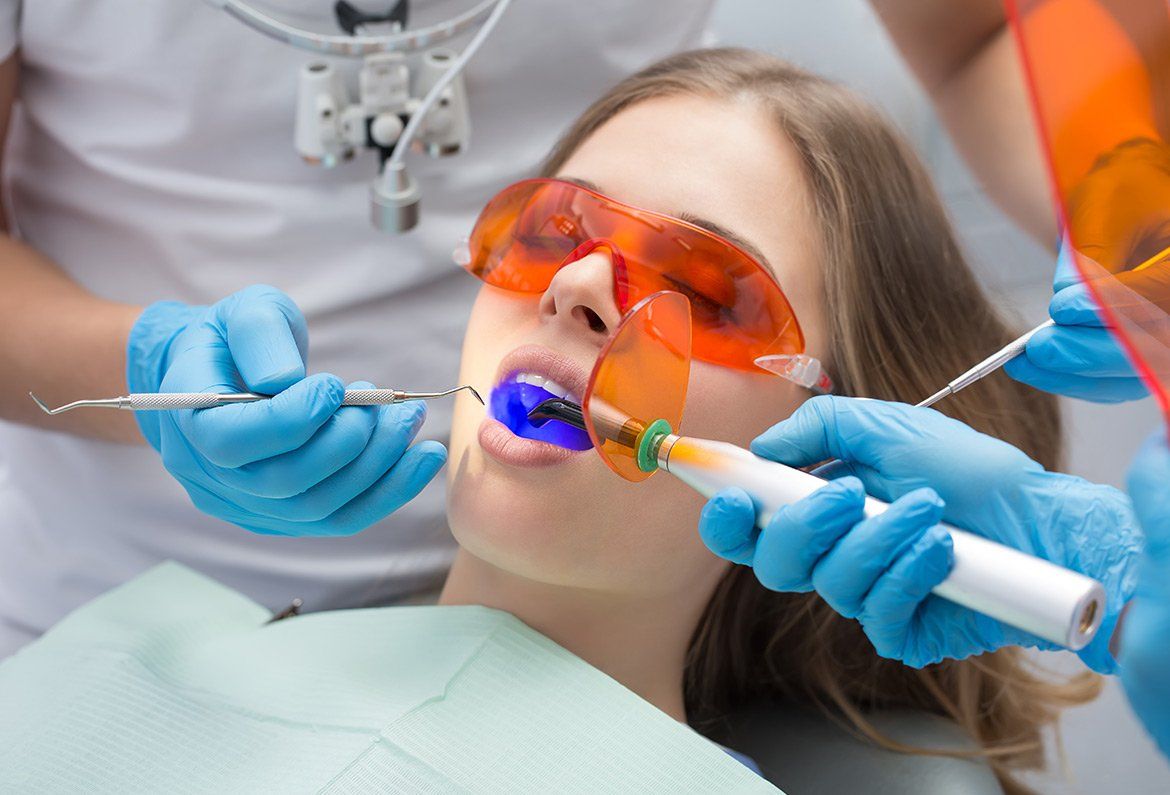 A dentist uses a blue dental curing light on a patient wearing orange protective glasses during a procedure.