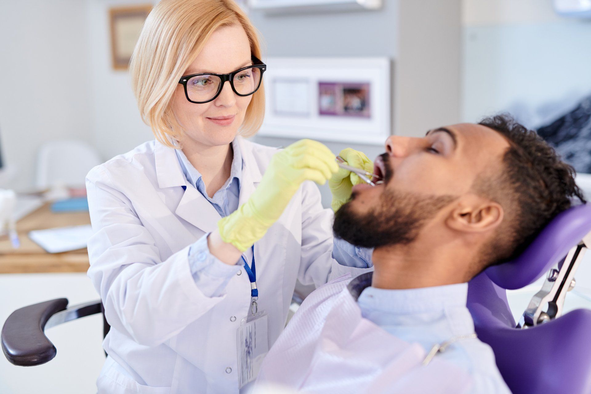 A dentist wearing a white coat and yellow gloves examines a patient's teeth in a dental chair.
