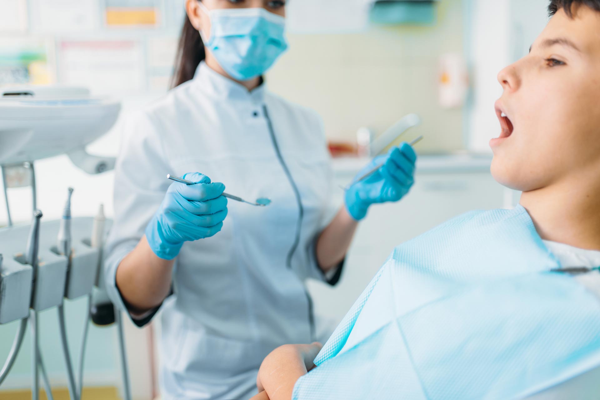 A dental professional in a mask and blue gloves holds tools while a patient sits in a chair with their mouth open.