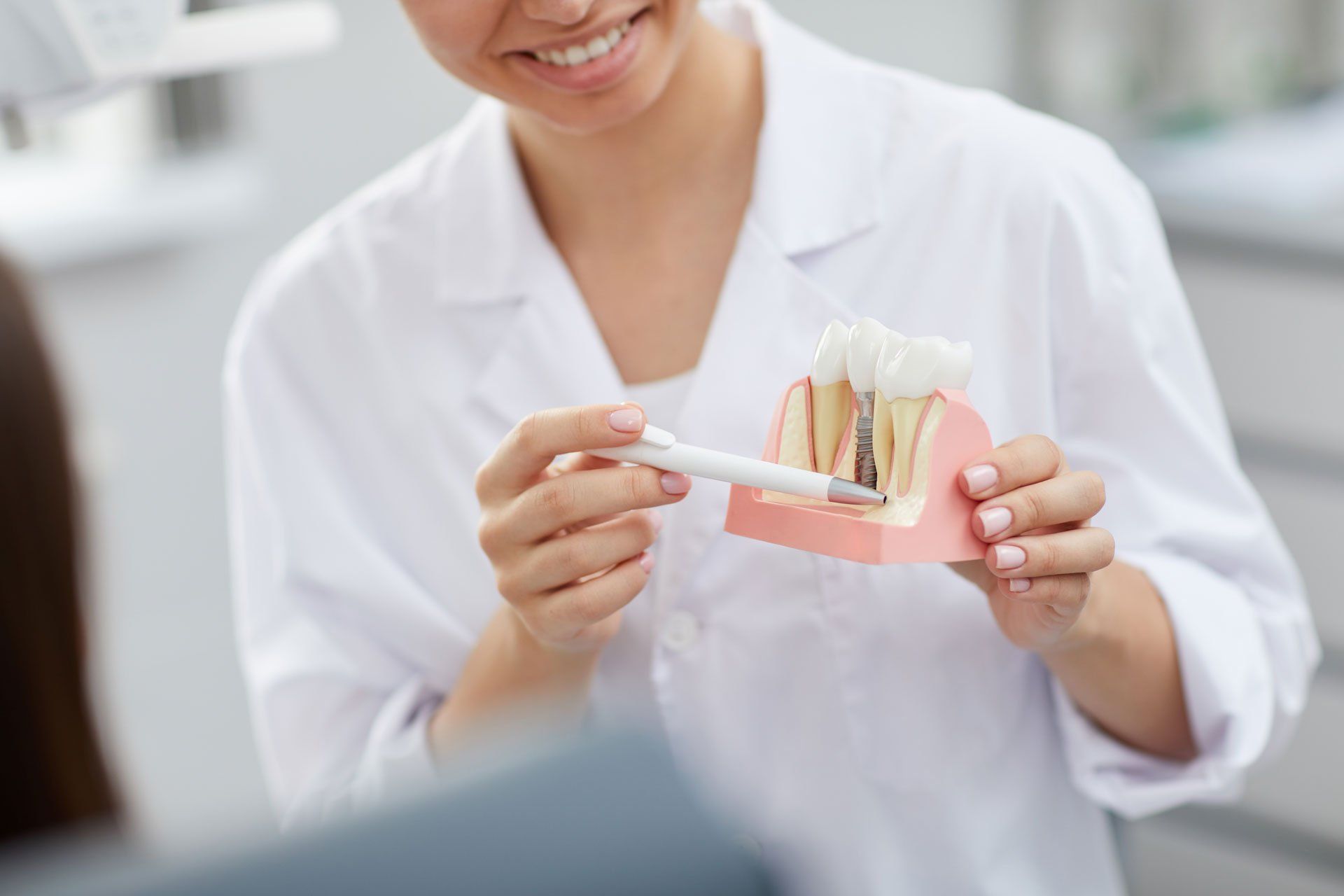 A dental professional in a white coat holds and points to a model of a dental implant, explaining it to a patient.