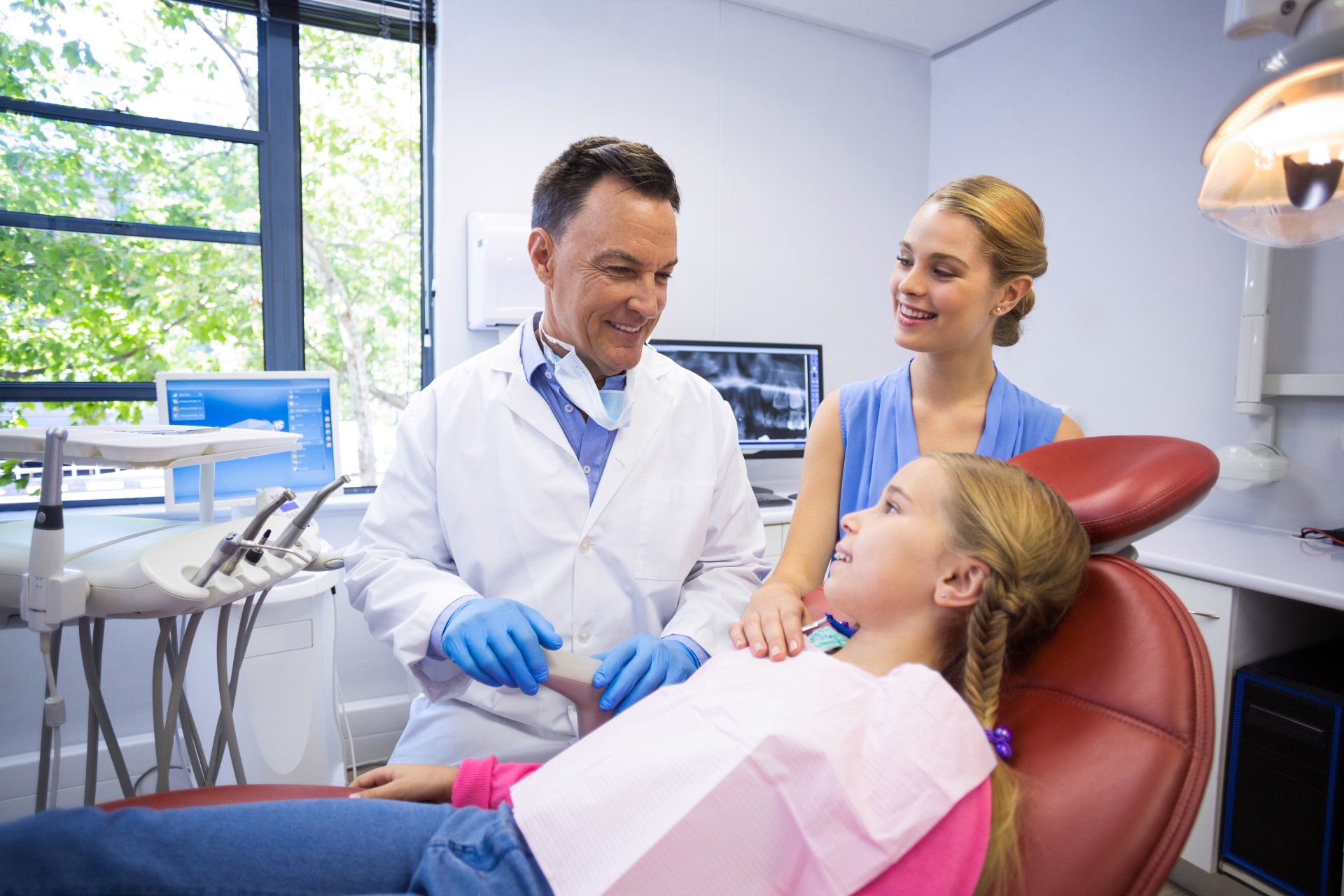 A dentist in a white coat talks to a child in a dental chair while an adult stands nearby in a bright, modern office.