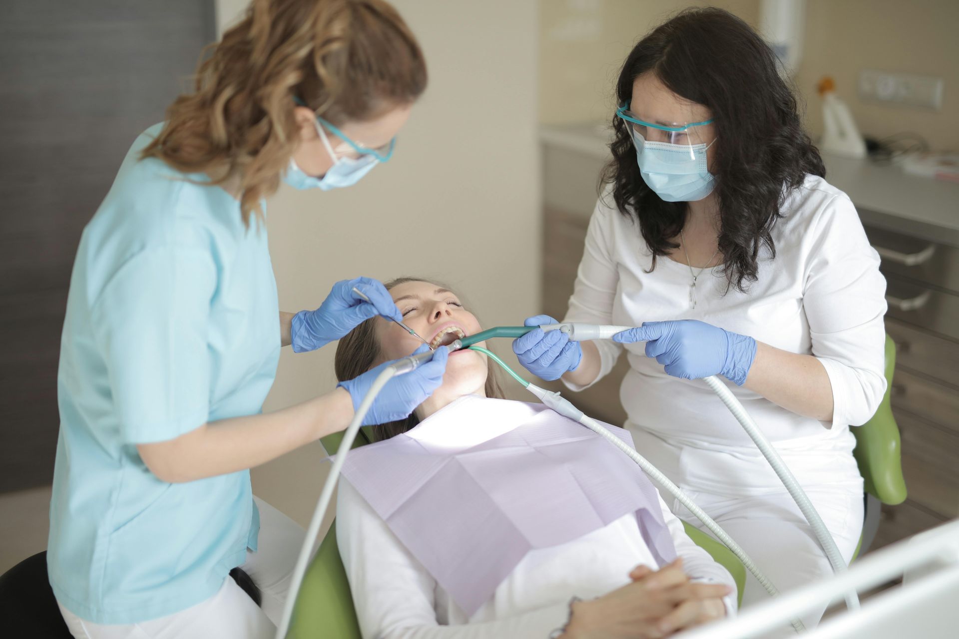 Two dental professionals in scrubs and masks perform a procedure on a patient in a dental chair.