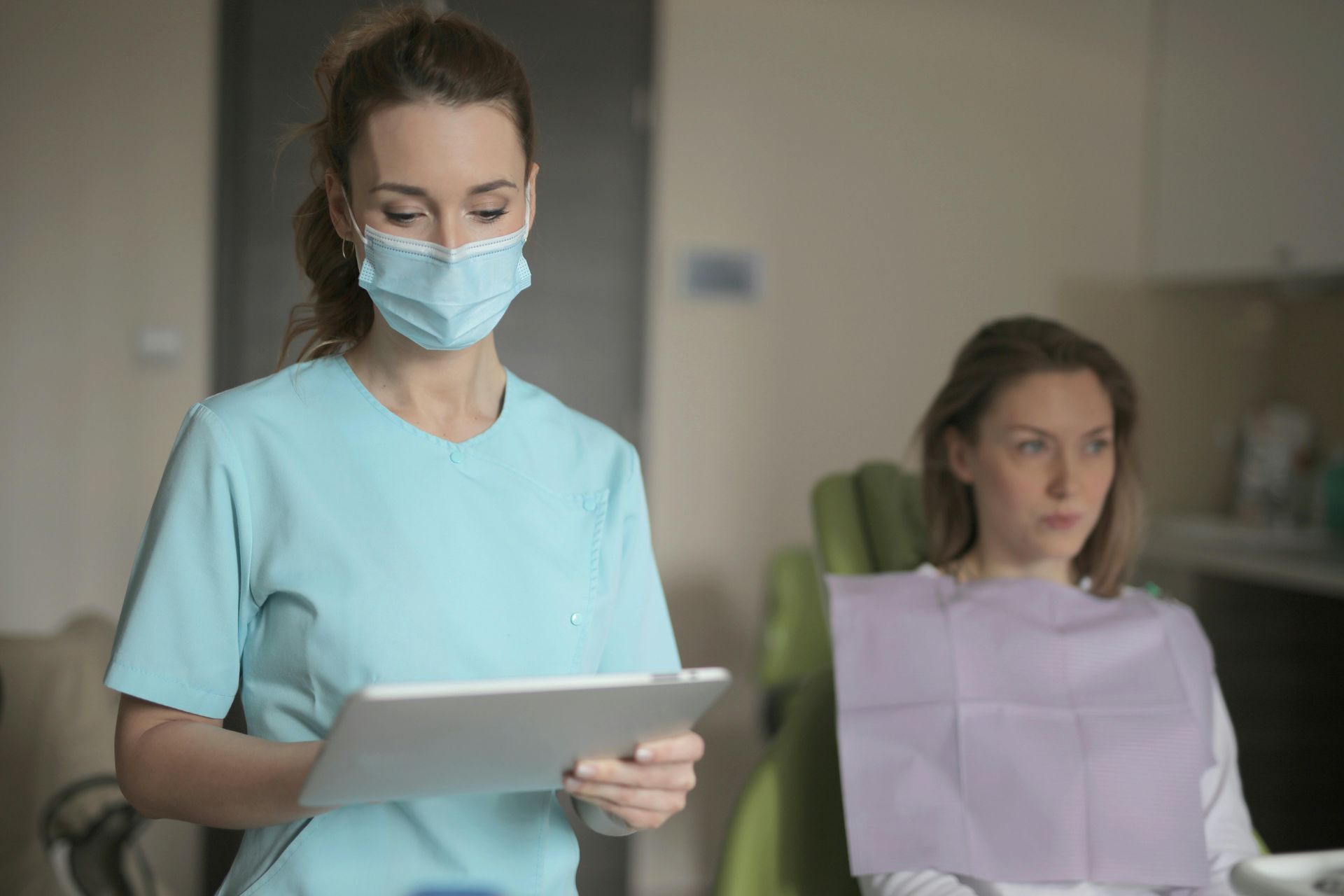 A dental professional in blue scrubs and a face mask holds a tablet while a patient sits in a dental chair nearby.