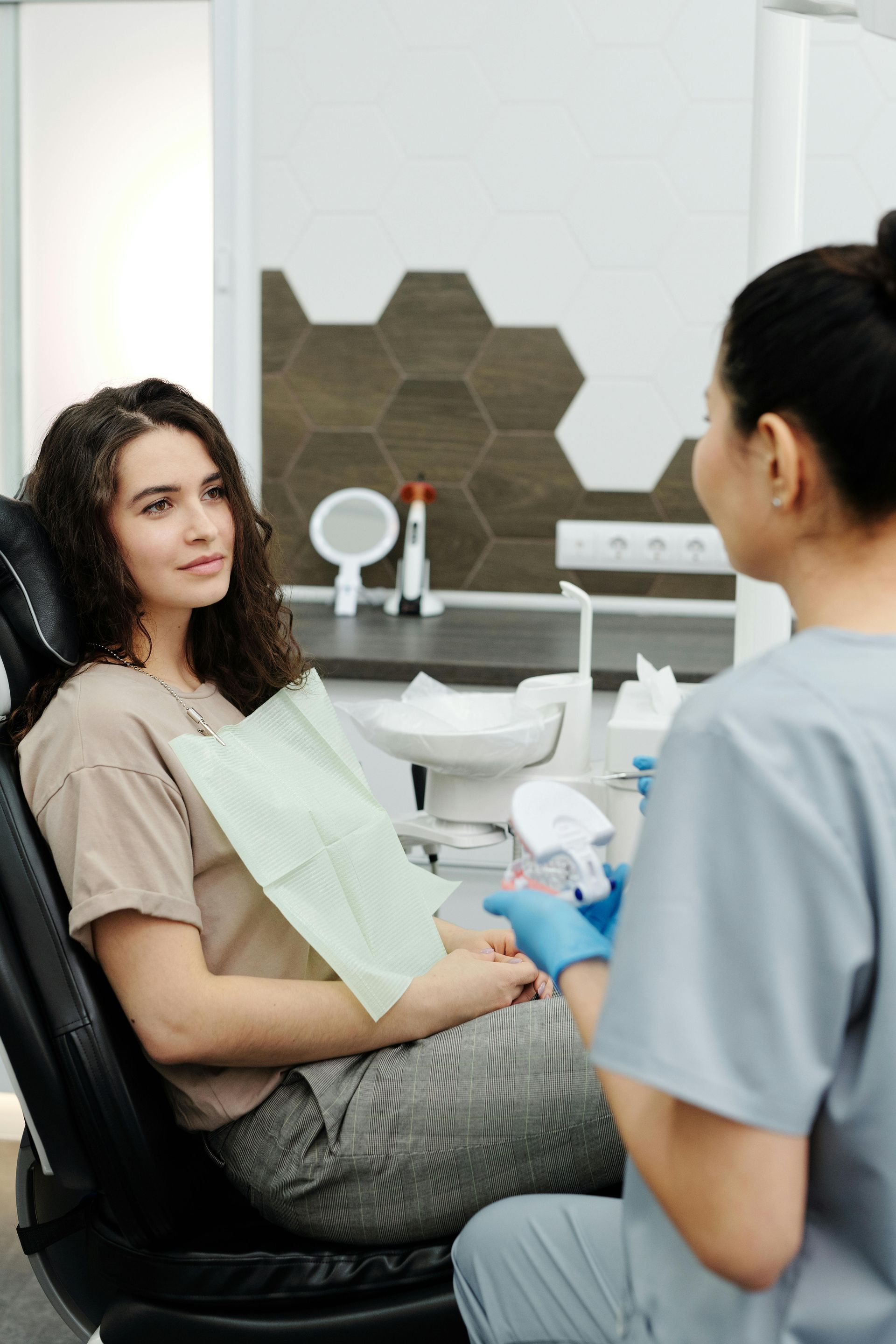 A patient wearing a dental bib sits in an exam chair, talking with a dental professional in a clinic.