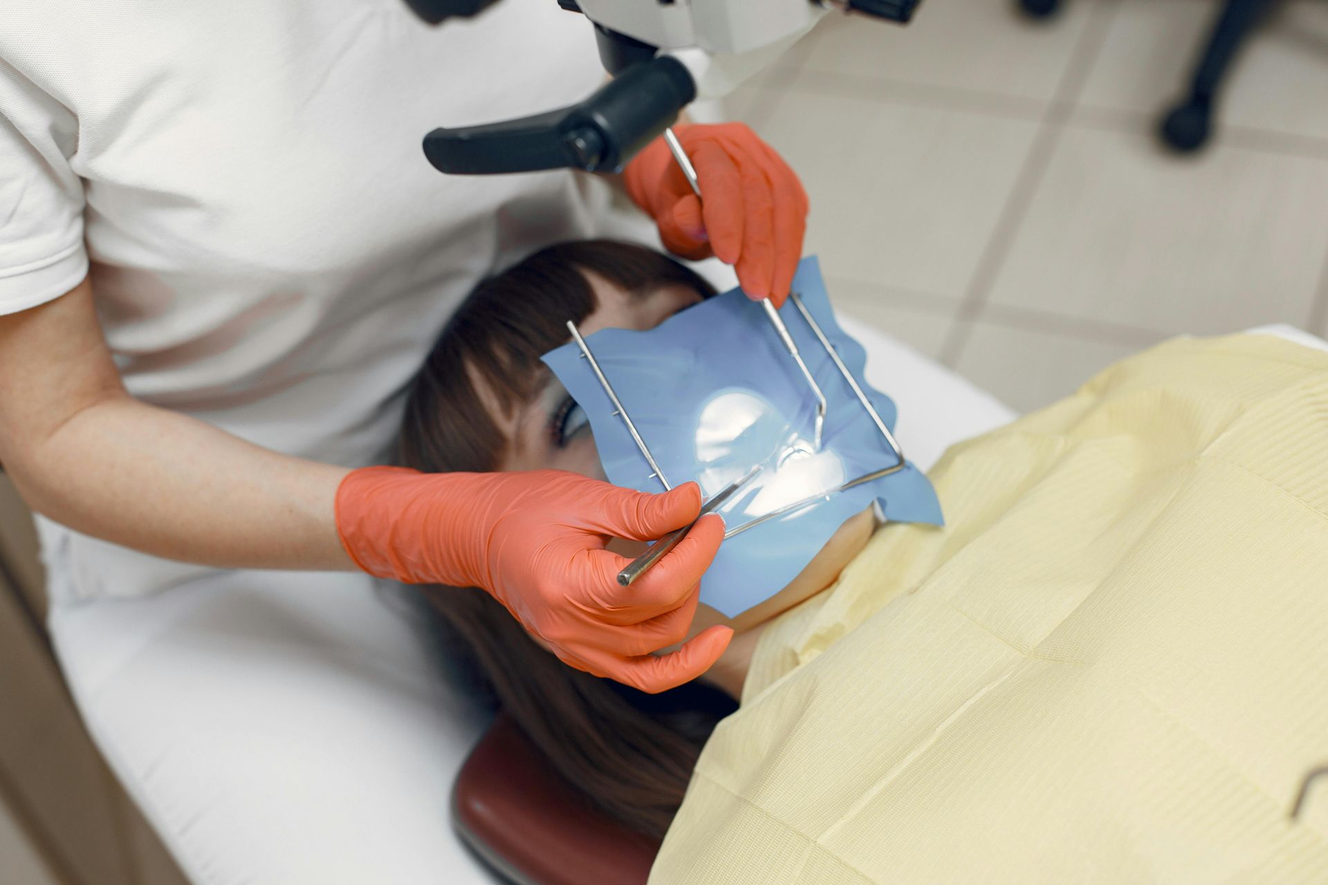 A dentist in orange gloves uses a dental mirror and tools while a patient undergoes treatment with a rubber dam.