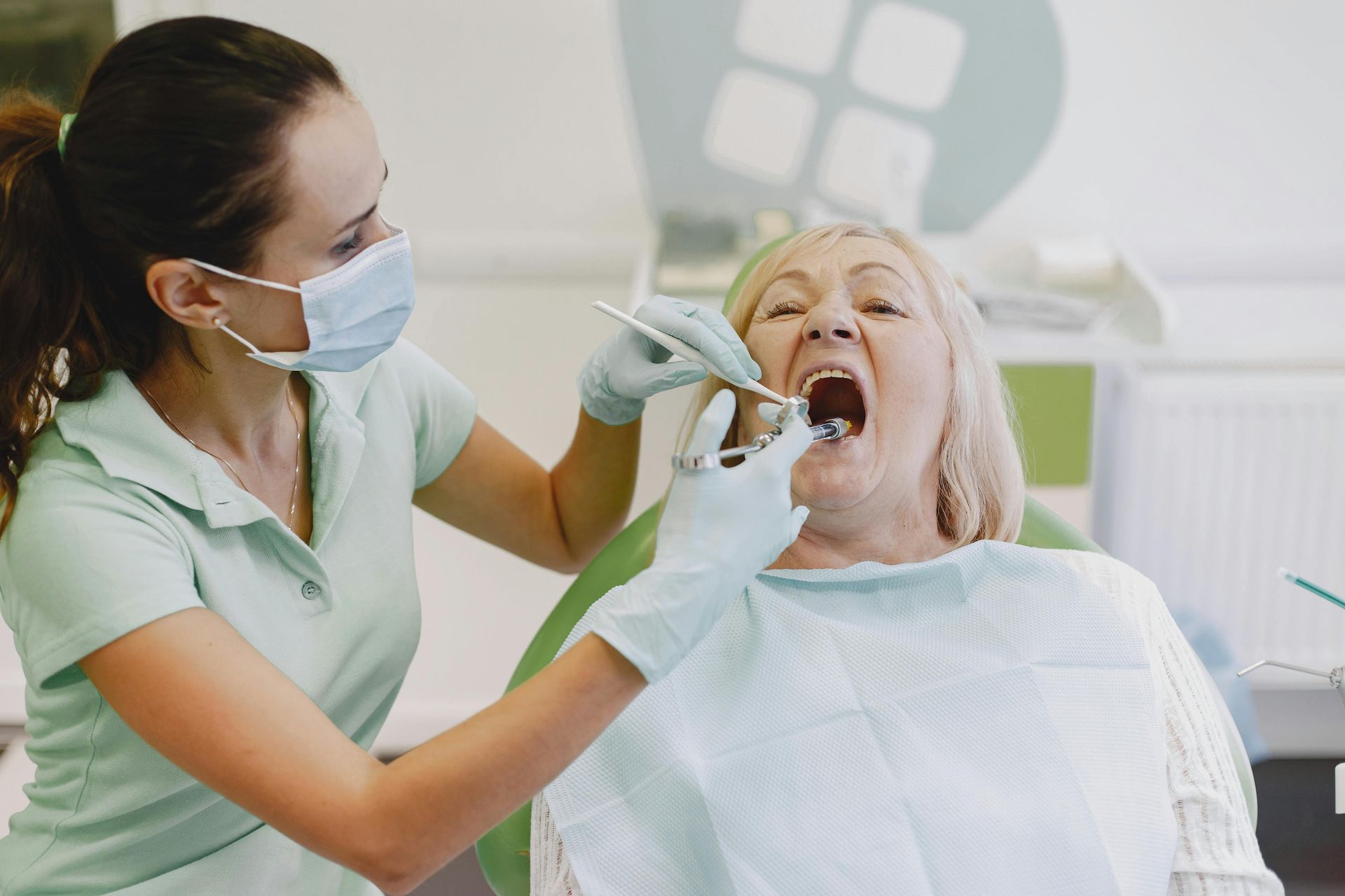 A dental professional in scrubs and a mask performing an examination on a patient in a dental chair.