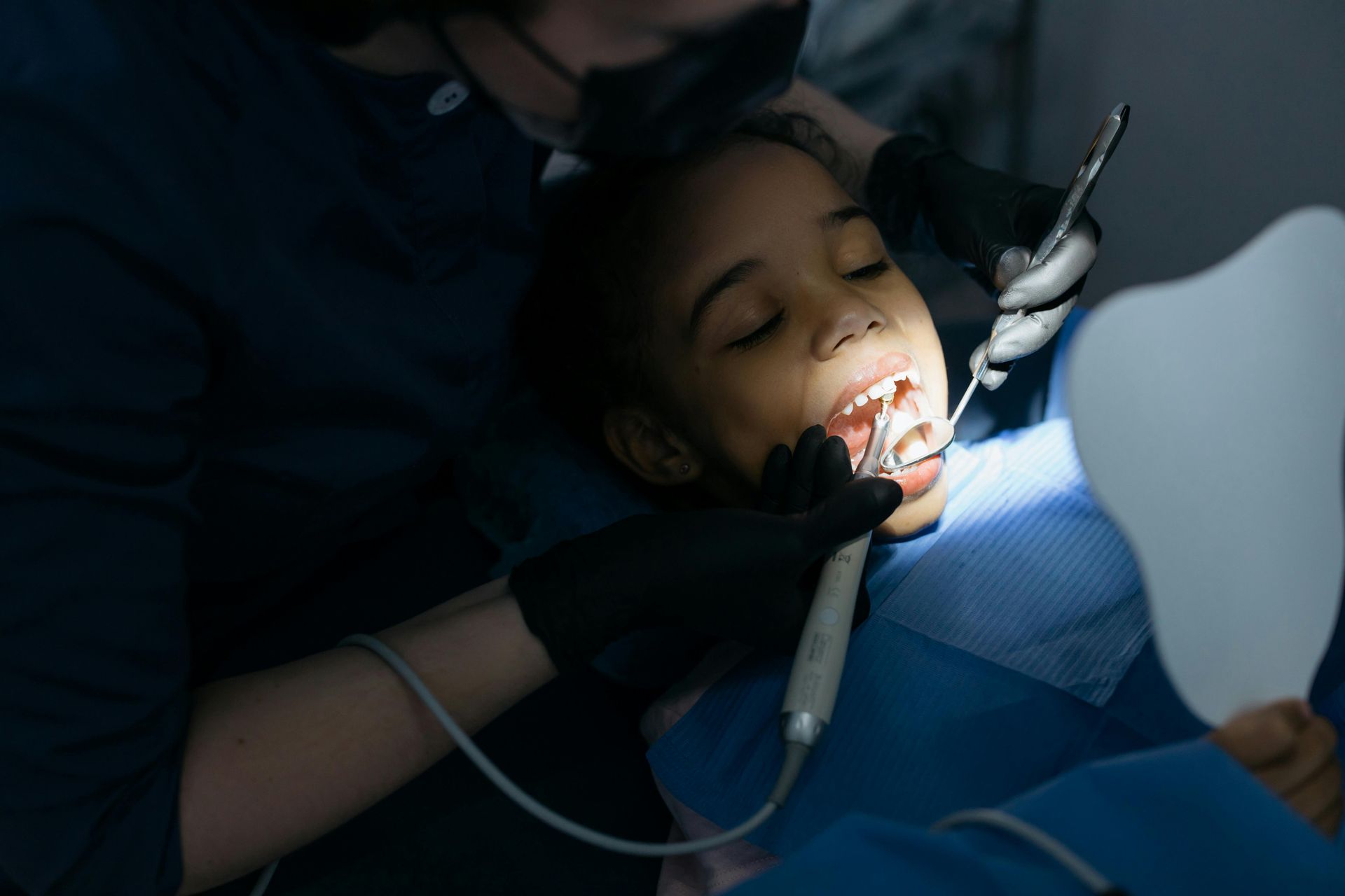 A dentist wearing black gloves examines a patient's teeth with dental instruments while the patient holds a mirror.