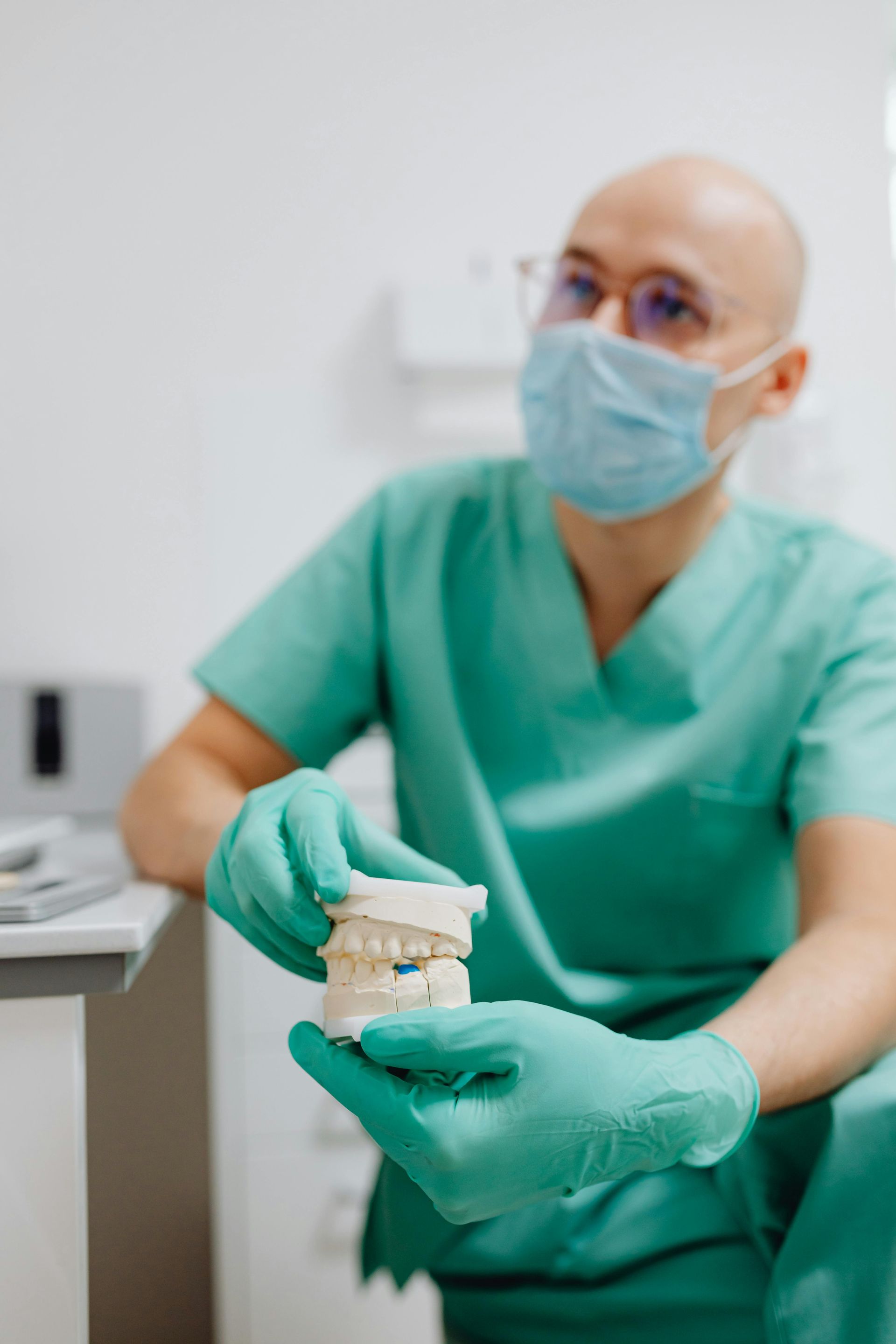 A dentist in green scrubs and a face mask holds a plaster dental model of teeth in a medical office.