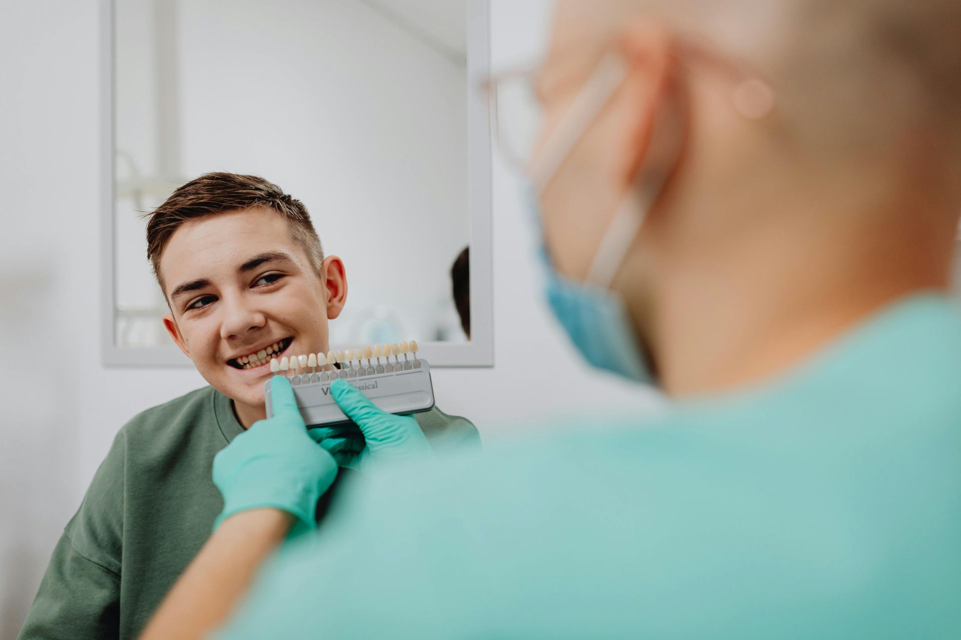 A dental professional in a mask and teal gloves holds a shade guide to a patient's teeth for a color match.