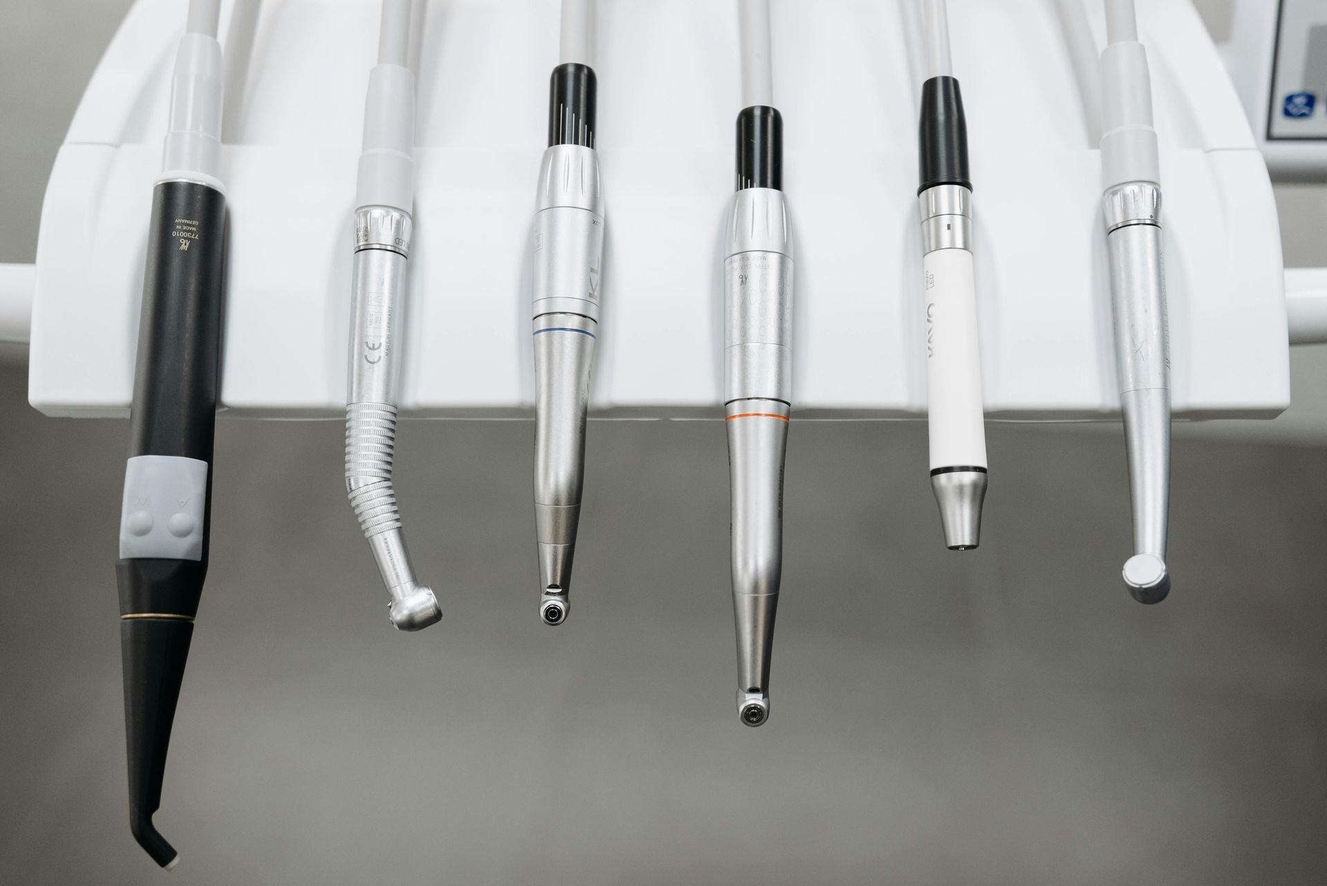 Six dental handpieces and tools hanging from a white bracket in a clinical setting.