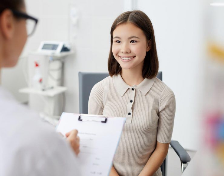 A person in a white coat holds a clipboard while speaking with a smiling individual in a medical office setting.