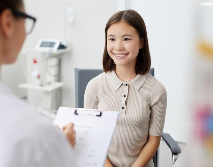 A person in a white coat holds a clipboard while consulting with a smiling individual sitting in an office.