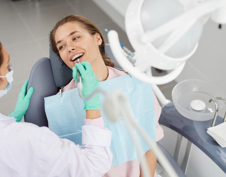 A person in a dental chair smiling while a dentist wearing gloves uses a dental tool to examine their teeth.