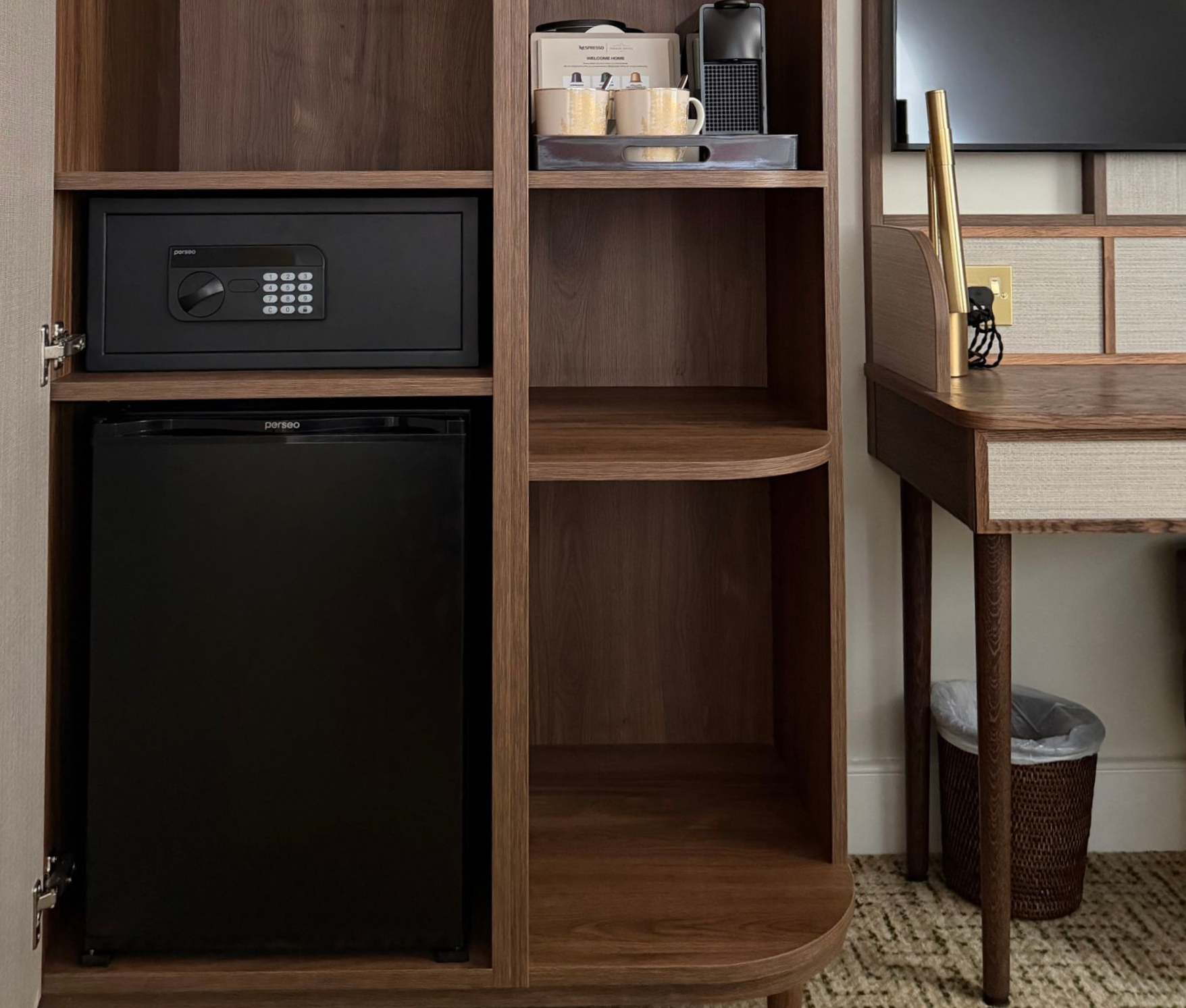 Wooden kitchenette with mini fridge, safe, kettle, shelving, and a small desk by a window and trash bin