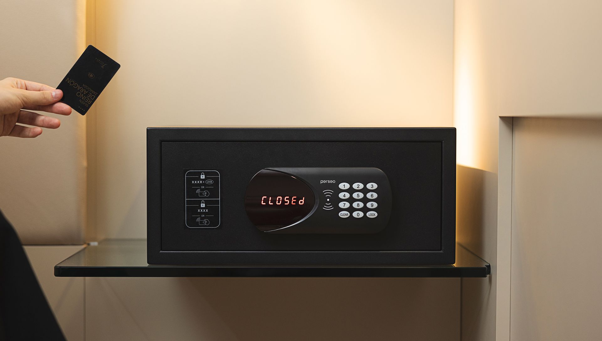 Hand holding a card near a black wall-mounted digital safe in a warmly lit room