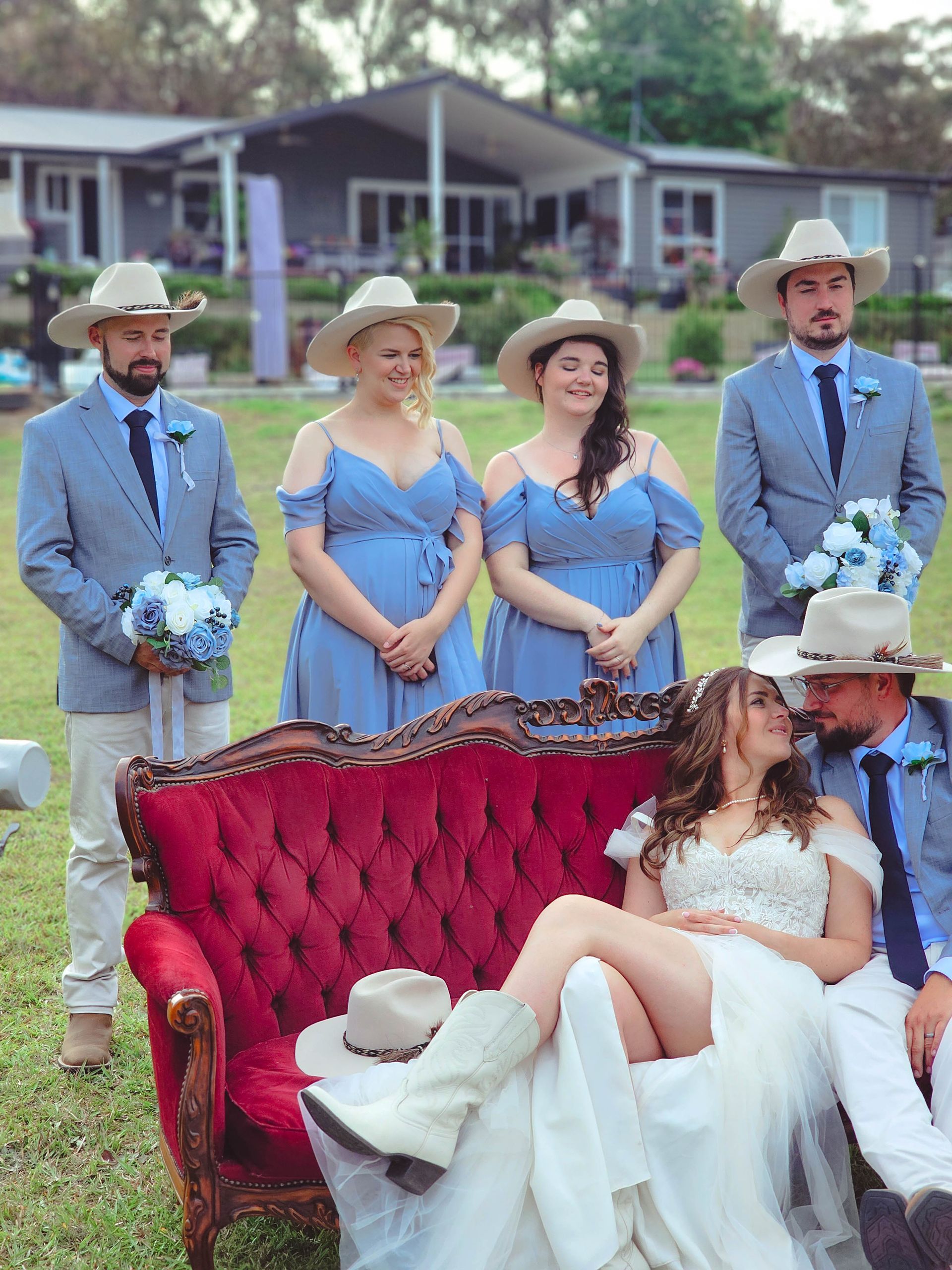 Wedding party posing outdoors beside a red couch, with a seated bride in white gown and attendants in blue.