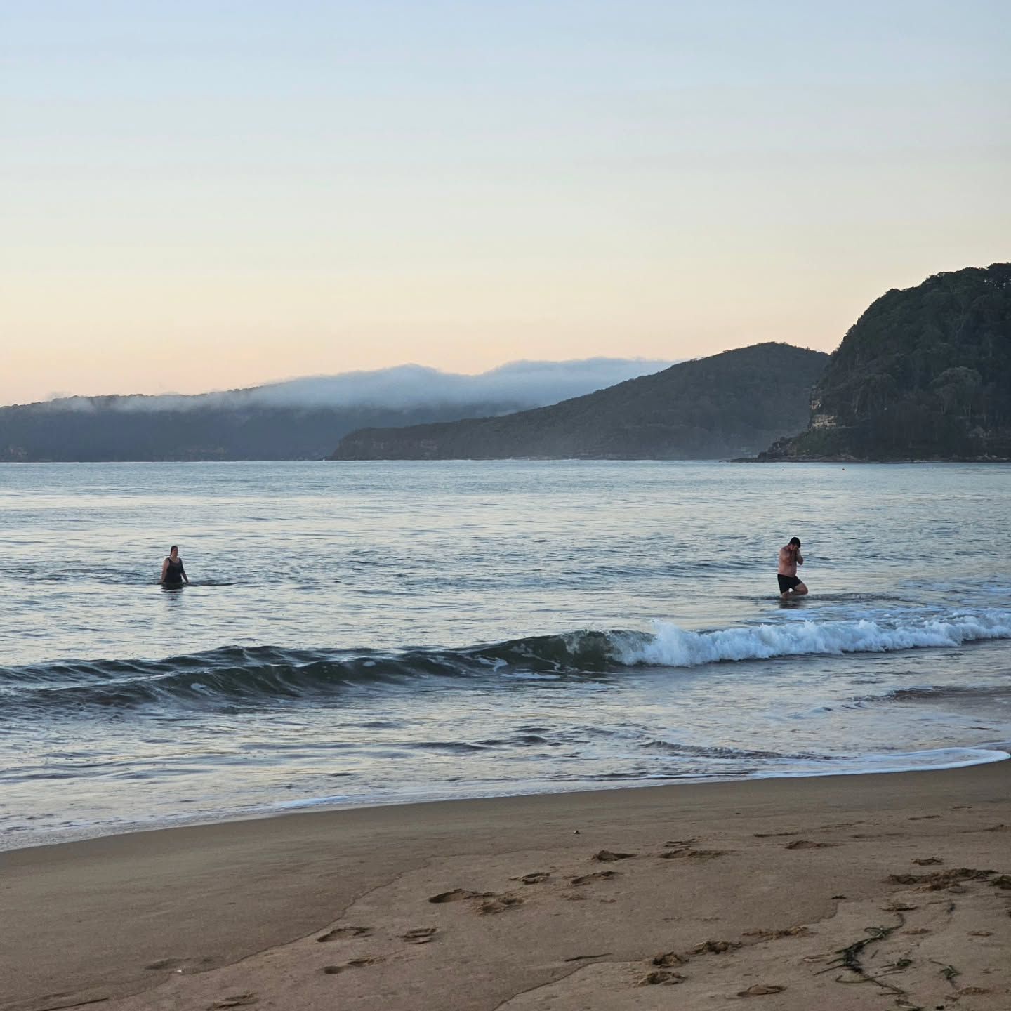 Two People Wade in Calm Ocean Waves at A Sandy Beach with Distant Hills at Sunset — Craig Stevens Photography in Pearl Beach, NSW