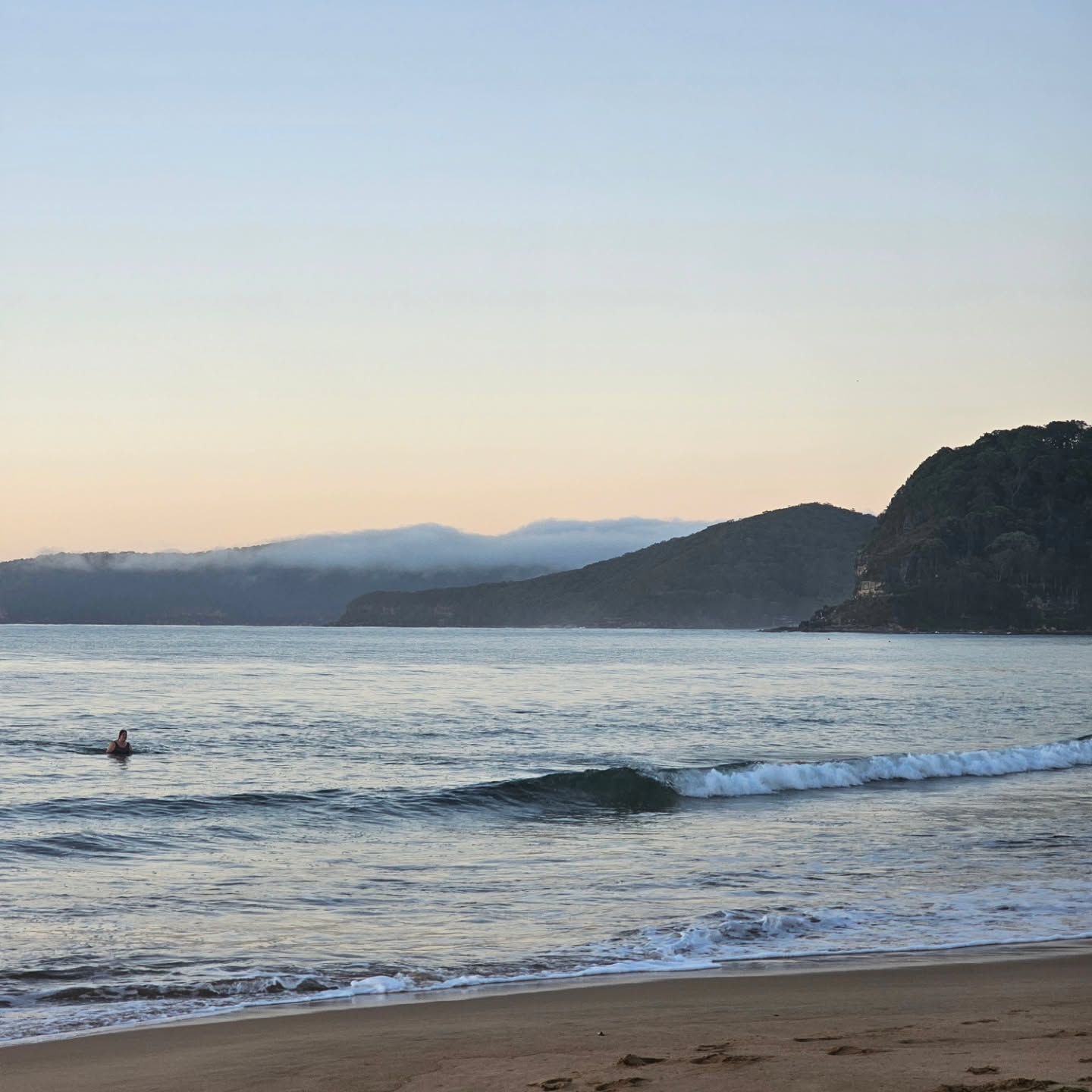 Calm Beach at Sunset with Small Waves, a Lone Surfer, and Distant Hills Under a Pastel Sky — Craig Stevens Photography in Pearl Beach, NSW