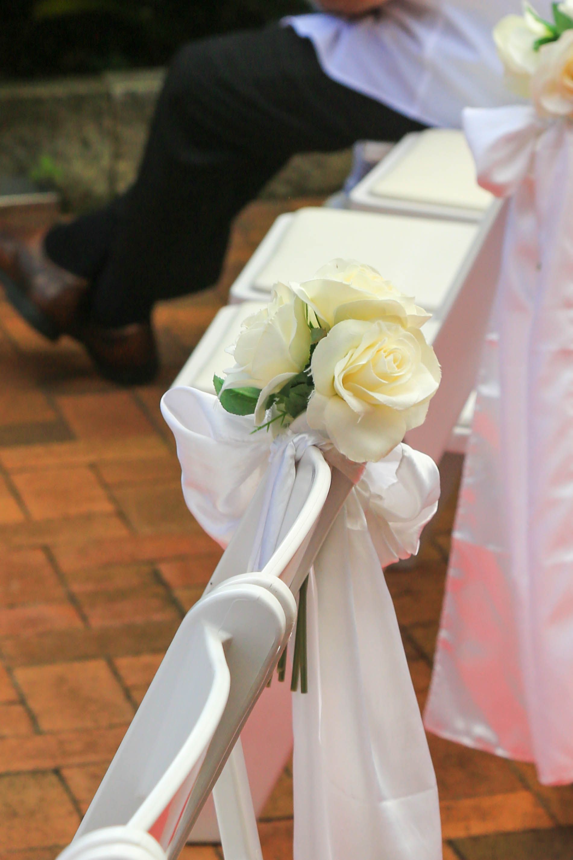 White Roses and Pink Ribbon Decorating a White Chair at An Outdoor Wedding Ceremony — Craig Stevens Photography in Pearl Beach, NSW