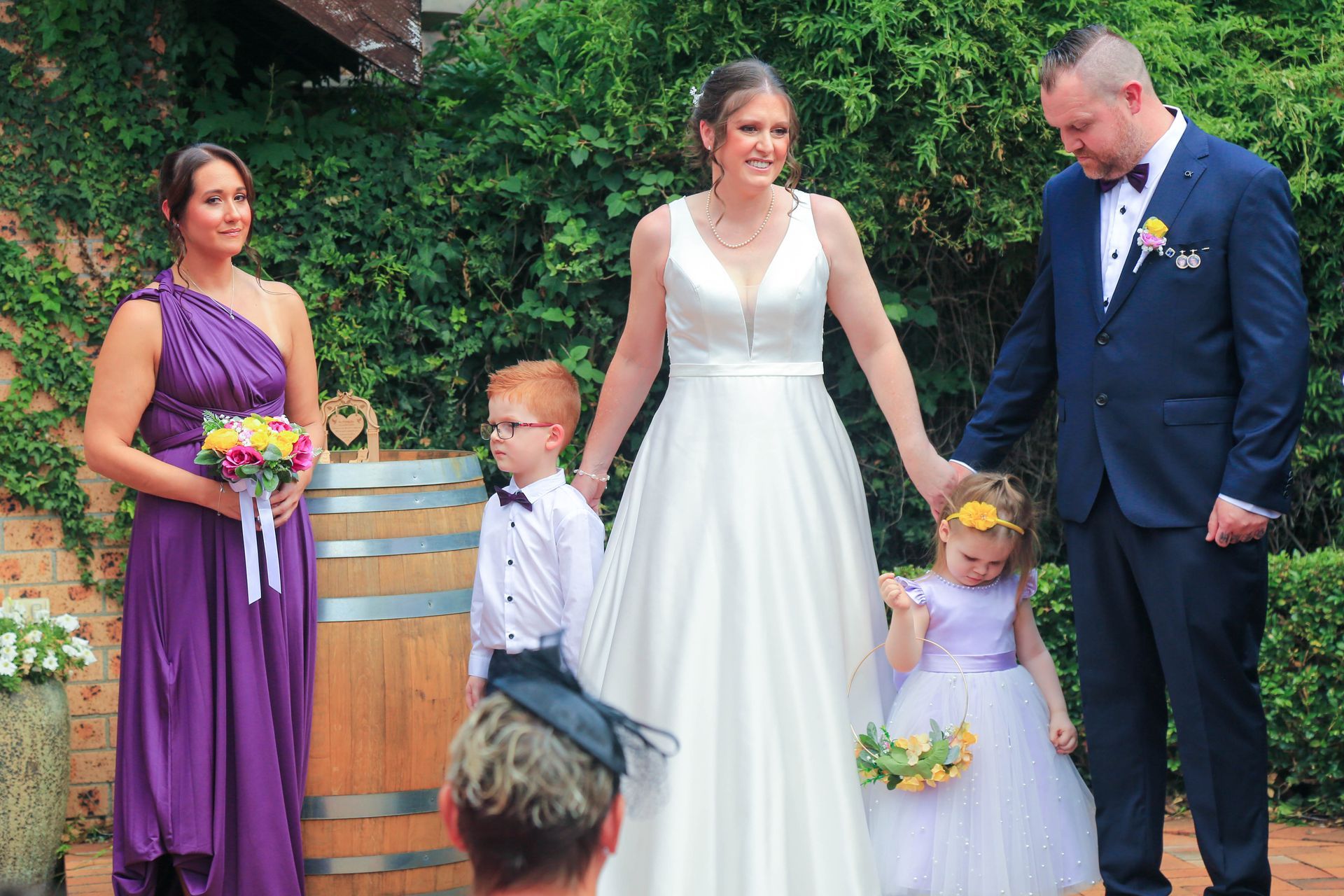 Wedding Party Standing Outdoors: Bride in White, Groom in Navy, Bridesmaids in Purple, Two Flower Girls — Craig Stevens Photography in Pearl Beach, NSW