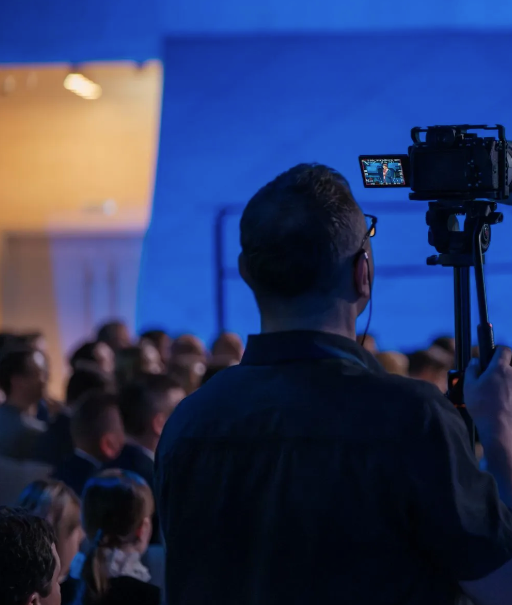 Audience seen from behind at a blue-lit event, with a camera filming on a tripod in the foreground