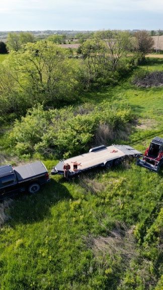 Two trucks towing a flatbed trailer in a grassy field near trees and open land.