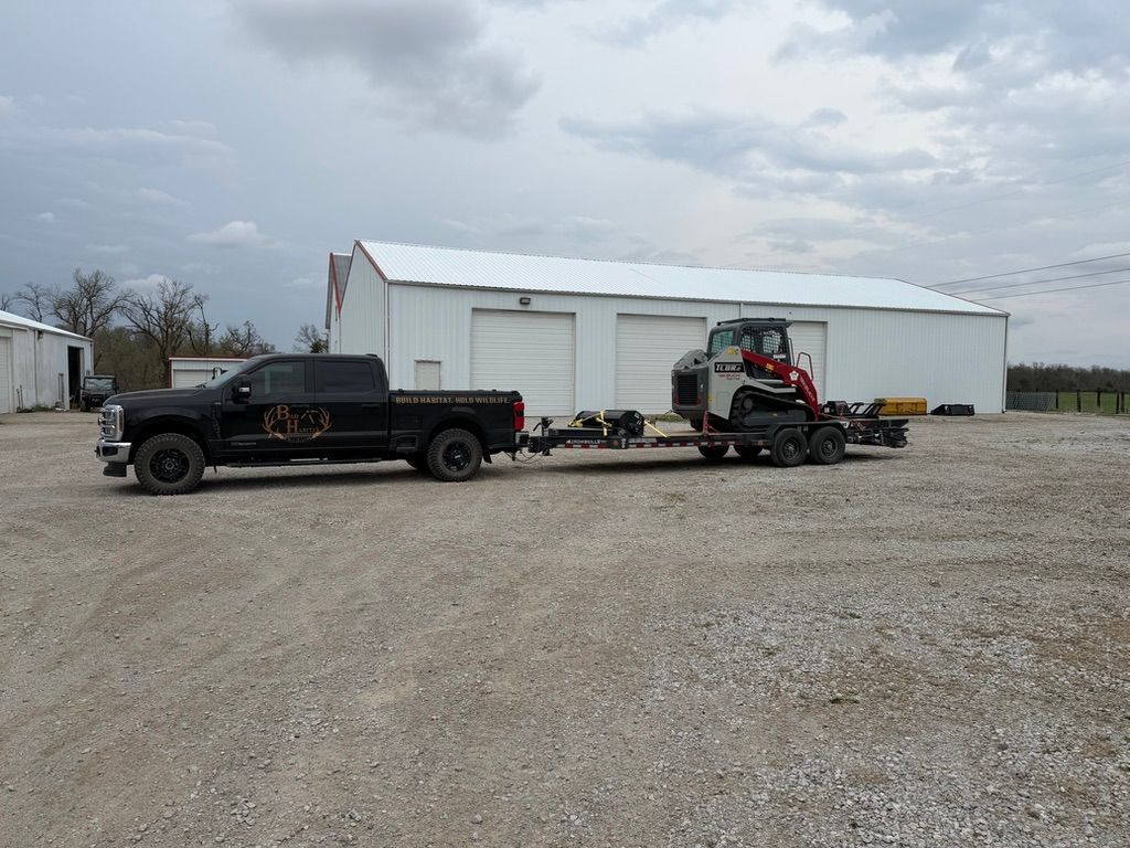 Black pickup truck towing a small skid steer beside a white metal building on a gravel lot