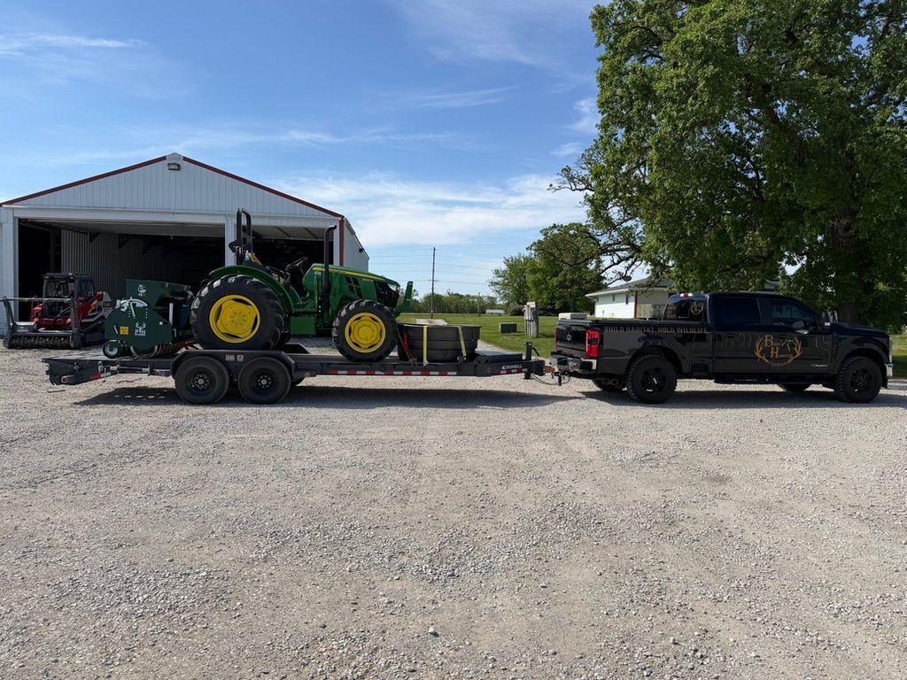 Green tractor on trailer beside black pickup in gravel lot near an open shed, under a blue sky