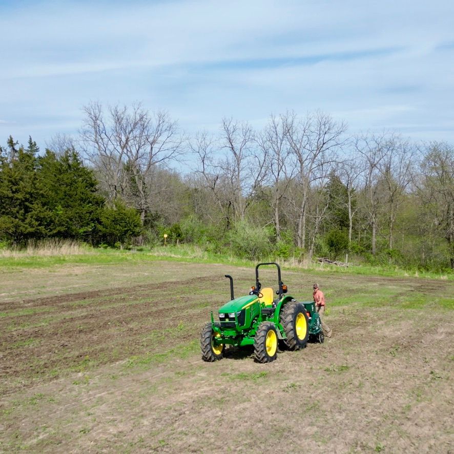 Green tractor in a field with a person standing beside it under a cloudy sky