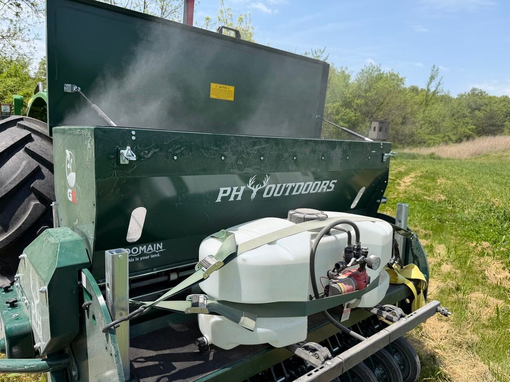 Green farm equipment with a white sprayer tank in a grassy field under a cloudy sky