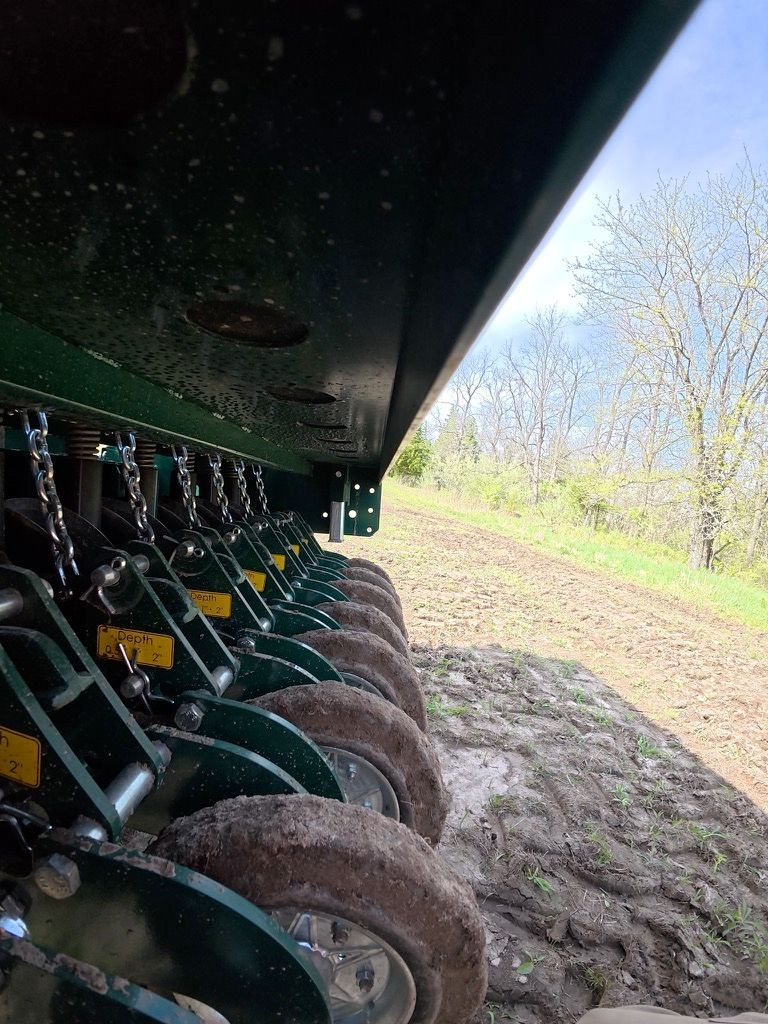 Rows of farm tillage discs under a green implement in a muddy field beside trees