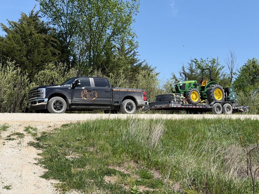 Black pickup truck towing a flatbed trailer with a green tractor on a rural roadside.