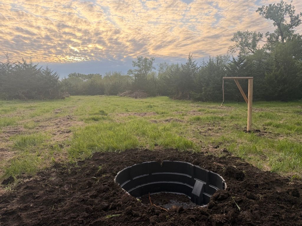 Grassy field at sunset with a black circular fire pit in the foreground and a wooden frame near the trees.
