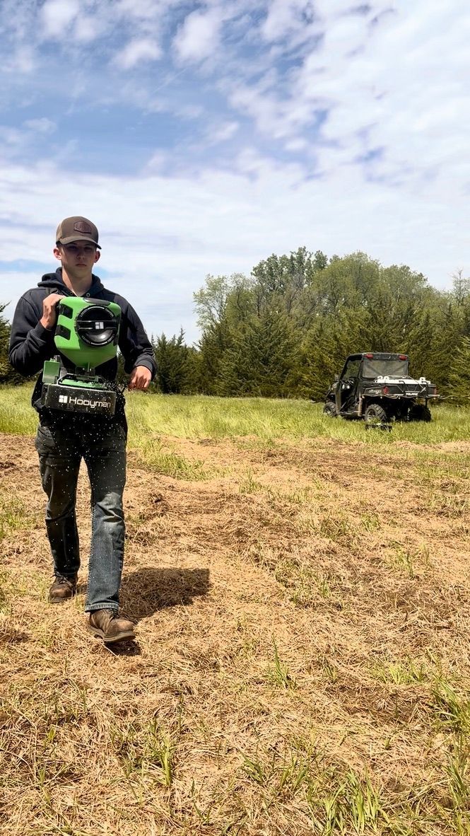 Person with green backpack walks across a grassy field; off-road vehicle sits in the background under a cloudy sky.